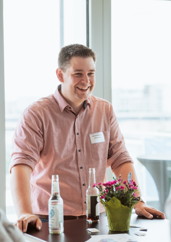 Ein lächelnder Mann in einem rosa Hemd steht an einem Tisch, während er mit anderen Personen spricht und eine entspannte Atmosphäre schafft.