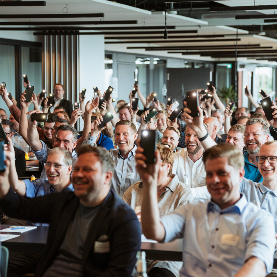 Eine große Gruppe von Menschen in einem Konferenzraum hält ihre Smartphones hoch. Die Atmosphäre ist begeistert und viele lächeln.