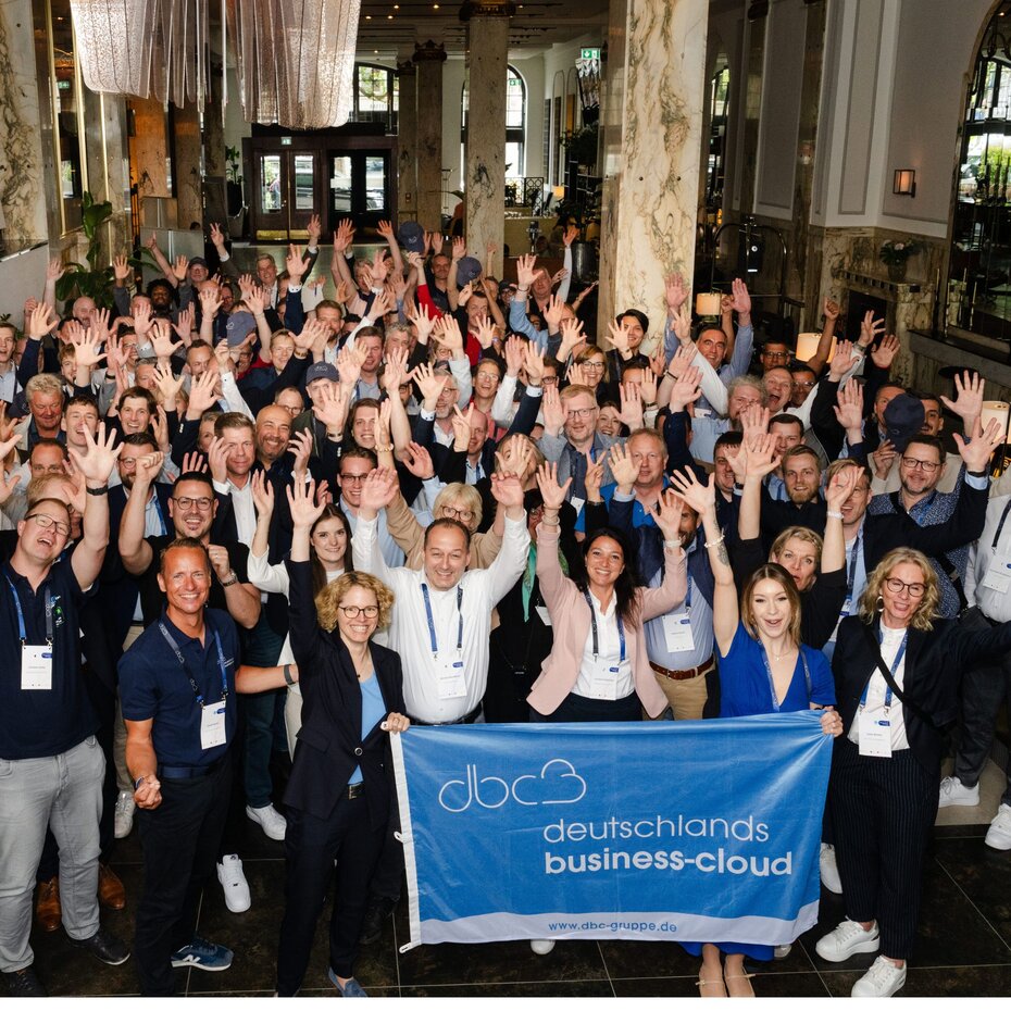 Eine große Gruppe von Menschen steht zusammen, lächelt und hebt die Hände. Im Vordergrund wird ein Banner mit der Aufschrift "Deutschlands Business-Cloud" gehalten.