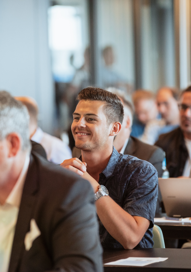 Eine Gruppe von Menschen hört aufmerksam bei einer Veranstaltung zu, während einige Teilnehmer lächeln und applaudieren.